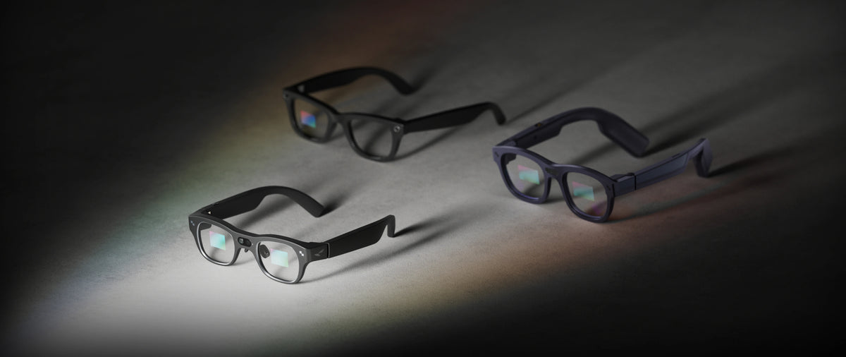 Three RayNeo X3 Pro glasses in black, gray, and navy colorways arranged on a textured surface under colorful lighting.