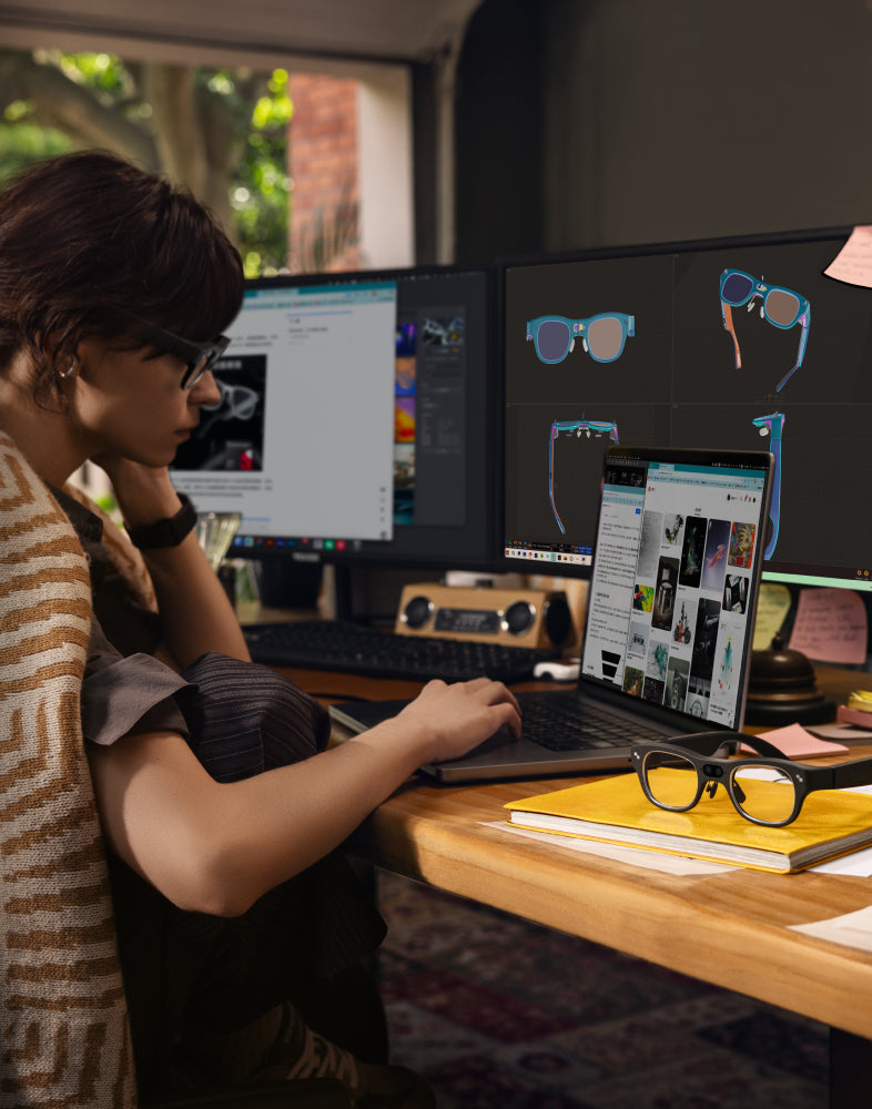 Woman working at a desk with dual monitors displaying smart-glasses design software and a pair of glasses resting on a nearby notebook.
